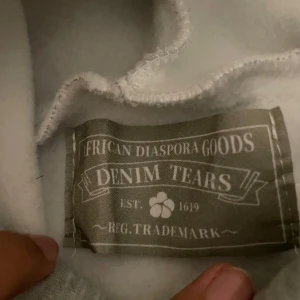 Grå hoodie från Denim Tears med blommor - Säljer en grå hoodie från Denim Tears med unika vita blommönster tryckta över hela tröjan. Hoodien har huva, långa ärmar och en klassisk känguruficka framtill. Perfekt för dig som vill sticka ut med en streetwear vibe.