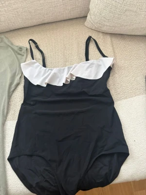 Svart baddräkt med volangdetalj - Snygg svart baddräkt med smala axelband och vit volang längs urringningen. Klassisk modell med enkel men trendig design, perfekt för stranden eller poolen. Materialet är stretchigt och följsamt för en bekväm passform.