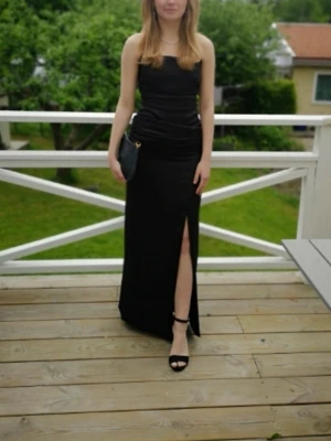Svart långklänning med slits - Elegant svart långklänning med hög slits framtill. Klänningen har en figurnära passform och är perfekt för speciella tillfällen. Matcha med klackar och en liten väska för en stilren look. Endast använd 1 gång! Jag är 163 