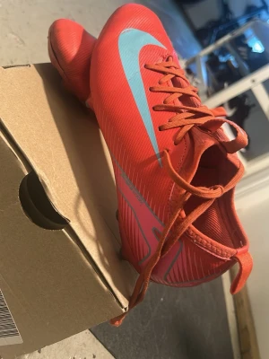 Röda Nike fotbollsskor med blå swoosh - Säljer ett par röda Nike fotbollsskor med blå swoosh och snörning. Skorna har en smal och sportig design med plastmaterial och mönstrade detaljer på sidorna. Perfekta för dig som vill ha stil och funktion på planen.
