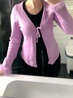 Ljuslila stickad cardigan med fluff - Ljuslila stickad cardigan med knytband framtill och fluffiga detaljer vid ärmsluten. Tröjan har en feminin v-ringning och är tillverkad i mjukt material som ger en skön känsla. Perfekt för dig som gillar trendiga och unika plagg.
