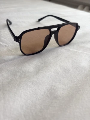 Svarta solglasögon - Coola solglasögon med svarta pilotbågar och bruna tonade glas. Bågarna har en klassisk form med dubbla nitar vid sidorna och en snygg detalj vid näsbryggan. Perfekta för att ge din look en extra edge under soliga dagar.