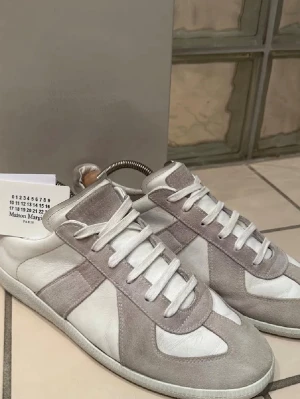  Äkta Maison Margiela gats   - Maison Margiela sneakers i en clean gråvit färgkombination med paneler i mocka och skinn. Klassisk rund tå, platt sula och snörning framtill. Snygg minimalistisk design som är lätt att matcha med olika stilar. Har kvar allt og som box , taggar, skopåsar, skohållare. Har även kvitto 😃 bara skriva, gärna byta mot andra gats med pengar till dig 🫵( är använda så finns tecken på slitage i men inget utanpå som stör ) se sista bild 🙌