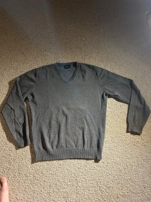 Grå V-ringad tröja från Boomerang - Klassisk grå stickad tröja från Boomerang med V-ringning och ribbade muddar vid ärmslut och nederkant. Tröjan är enkel och stilren, perfekt att matcha med skjorta eller t-shirt. Materialet känns mjukt och bekvämt.