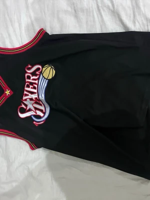 Svart Sixers basketlinne - Säljer ett svart basketlinne med Philadelphia 76ers-logga framtill. Linnet har röda och vita detaljer runt halsringning och ärmhål samt ett broderat märke med en gul basketboll. Perfekt för basketträning eller streetwear.
