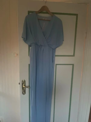 Ljusblå omlottklänning lång - Långklänning i ljusblått med omlott-design och korta ärmar. Klänningen har ett flowigt fall och markerad midja, perfekt för dig som gillar en elegant och enkel stil. Materialet känns lätt och bekvämt mot huden.      . Ny med prislapp men dragkedjan har gått sönder troligen lätt att laga 