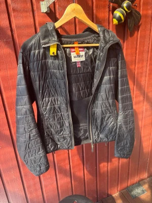 Svart pufferjacka från Parajumpers - Säljer en svart pufferjacka från Parajumpers med huva och dragkedja framtill. Jackan har quiltad design, meshpaneler på sidor och rygg samt en gul detalj vid halsen. Märkespatch på ärmen och praktiska fickor. Perfekt för kalla dagar.
