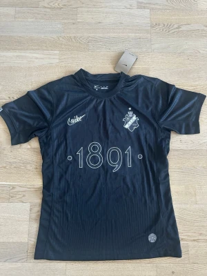 Svart AIK tröja från Nike - Säljer en svart AIK tröja från Nike med tryck '1891' på framsidan och AIK:s klubbmärke. Tröjan är tillverkad i Dri-FIT material som är perfekt för sport och träning. Rund halsringning och korta ärmar. Snygg och stilren design för AIK-fans.