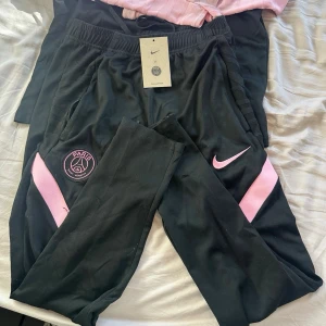 Svarta Nike PSG träningsbyxor - Svarta träningsbyxor perfekt för sport och träning. Snyggt rosa Nike-märke på höften och rosa paneler längs benen.