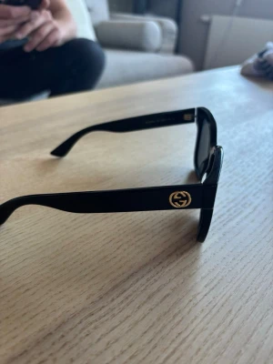 Gucci glasögon dam - Snygga svarta solglasögon från Gucci med klassisk fyrkantig form och mörka linser. Bågarna är i blankt syntetmaterial och har Gucci-loggan i guld på sidan. Perfekt för dig som vill ha en stilren och lyxig look.