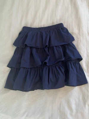 Marinblå volangkjol med resår - Säljer en marinblå kjol med flera lager volanger och elastisk midja. Kjolen har en luftig och flowy känsla, perfekt för dig som gillar att sticka ut med en cool och ungdomlig stil. Materialet känns som bomull och kjolen är kort.