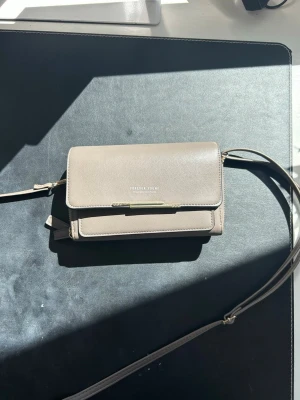 Grå axelväska från Forever Yours - Snygg grå axelväska från Forever Young med stilren design och guldfärgade detaljer. Väskan har ett lock med magnetknäppning, flera fack med dragkedja och en justerbar axelrem. 