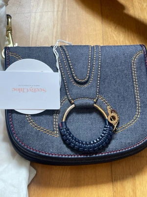 Chloé denim handväska med ringdetalj - Snygg handväska från Chloé i mörkblå denim med kontrasterande gul söm och röda detaljer. Väskan har en unik rund ring i metall och läder på framsidan, samt en magnetisk stängning. Perfekt för dig som vill ha en trendig och stilren accessoar.