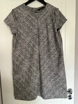 Kortärmad mönstrad klänning från Zara - Säljer en snygg kortärmad klänning från Zara Basic Collection. Klänningen är vit med svart mönster som liknar små blad eller fjädrar och har en rak, enkel form. Perfekt för dig som gillar stilrena och grafiska plagg.