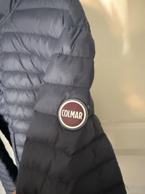  Colmar jacka - Säljer en mörkblå dunjacka från Colmar med huva och två fickor med dragkedja. Jackan har ett quiltat mönster och Colmar-loggan på ärmen. 