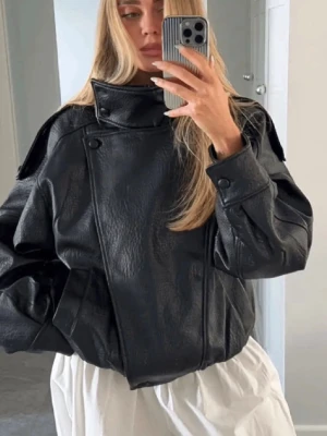 Svart oversized skinnjacka - Cool svart skinnjacka med hög krage och asymmetrisk knäppning. Jackan har en oversized passform, breda axlar och detaljerade sömmar. Perfekt för dig som vill ha en edgy och trendig look. Passar till jeans eller kjol för en modern stil.