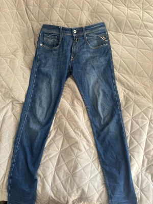 Blå jeans från Replay - Snygga blå jeans från Replay med smal passform och klassisk femficksdesign. Jeansen har ljusa slitningar framtill och bakfickor med dekorativa sömmar. Tillverkade i denim med en bekväm känsla och detaljer som metallknapp och läderpatch bak.