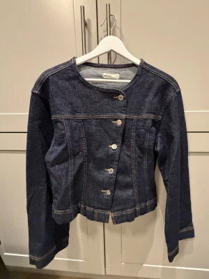 Mörkblå jeansjacka med sned knäppning - Snygg mörkblå jeansjacka från Gina Tricot med unik sned knäppning framtill. Jackan har rund halsringning, långa ärmar och kontrastsömmar. Perfekt för dig som vill ha en klassisk men ändå annorlunda jeansjacka i garderoben.