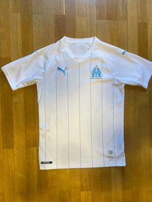 Vit Marseille matchtröja från Puma - Snygg vit matchtröja från Puma med Olympique de Marseille-logga och tunna blå ränder. Tröjan har korta ärmar, V-ringning och är tillverkad i ett lätt, andningsbart funktionsmaterial. Perfekt för fotbollsträning eller att bära till vardags.