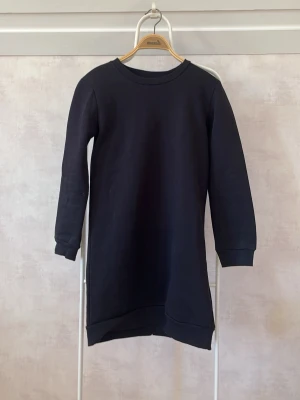 Svart sweatshirtklänning - En enkel och stilren svart sweatshirtklänning med rund halsringning och långa ärmar. Klänningen har en rak passform och är tillverkad i ett mjukt sweatshirtmaterial som känns bekvämt mot huden.