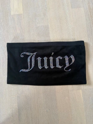 Svart bandeau tubtopp Juicy Couture - Säljer en svart bandeau tubtopp från Juicy Couture med glittrande stenar som stavar 'Juicy' framtill. Toppen är axelbandslös och har en mjuk, stretchig känsla. Perfekt för dig som vill ha en statement piece i garderoben.