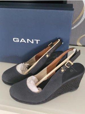 Gant kilklack-sandaler  - Snygga svarta sandaler från GANT med kilklack och justerbar rem med guldfärgat spänne. Ovandel i textil och klacken är täckt med flätad jute för en cool look. Perfekta för dig som vill ha både stil och komfort.