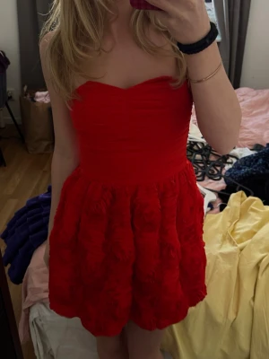 Röd axelbandslös kortklänning - Axelbandslös röd klänning med strukturerad och voluminös kjol med rosformade detaljer. Överdelen är draperad och figurnära, vilket ger en snygg och modern siluett. Perfekt kräftskivan❤️