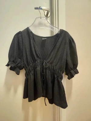 Svart blus med snörning från Gina Tricot - Säljer en svart blus från Gina Tricot med puffiga korta ärmar och v-ringning. Blusen har snörning i midjan och är tillverkad i ett strukturerat syntetmaterial. Perfekt för dig som gillar en feminin och trendig look.