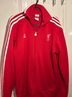 Röd Liverpool Adidas träningsjacka (2011-12) - Röd träningsjacka från Adidas med Liverpool FC-logga broderad på bröstet. Jackan har vita ränder längs axlarna och ärmarna, hög krage och dragkedja framtill. Materialet är syntetiskt och jackan har två sidofickor. Perfekt för fotbollsfans och sportiga dagar.