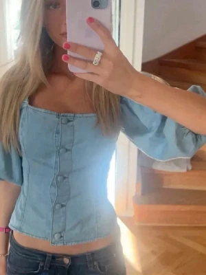 Ljusblå korsettopp med puffärmar - Snygg ljusblå korsettopp med puffiga kortärmar och knappar framtill. Toppen har en fyrkantig halsringning och är croppad, vilket ger en trendig look. Perfekt för dig som vill sticka ut med en cool denimstil.