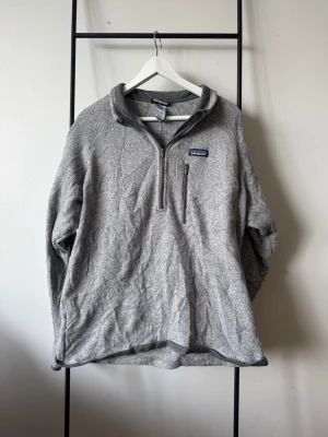 Grå fleece-tröja från Patagonia - Mjuk och varm grå fleece-tröja från Patagonia med halv dragkedja och bröstficka med zip. Tröjan har hög krage och logotyp på bröstet. Perfekt för lager-på-lager och chill dagar. Materialet är fleece och modellen är långärmad.