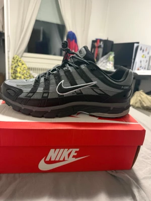 Nike P-6000 svart/grå sneaker - Nike P-6000 sneaker i svart och grå mesh med syntetiska detaljer och vit swoosh på sidan. Skon har en sportig design med robust sula och klassiska snörningar. Perfekt för dig som gillar en clean och modern look.