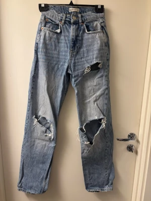 Ljusblå slitna jeans från Gina tricot  - Säljer ett par ljusblå jeans från Gina tricot med flera stora slitningar och hål på benen och bakfickorna. Jeansen har klassisk femficksdesign, raka ben och hög midja. Perfekta för dig som gillar en avslappnad och trendig look.