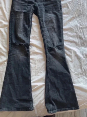 Mörkgrå bootcut jeans med slitningar - Snygga mörkgrå bootcut jeans med slitna detaljer vid knäna och stora fickor framtill. Jeansen har en utsvängd passform och är perfekta för en avslappnad och trendig look. Materialet är klassisk denim.