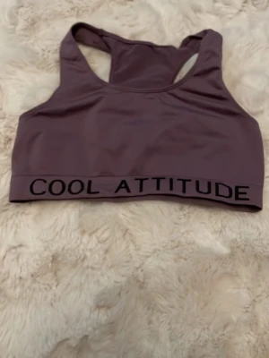 Lila sport-bh med text COOL ATTITUDE - Sport-bh i lila färg med bred resår och texten COOL ATTITUDE framtill. Modellen har brottarrygg och är tillverkad i ett stretchigt material som passar perfekt för träning och aktivitet. Strorlek 158/164, pris 10kr 