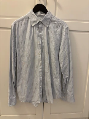 Randig skjorta från These Glory Days - Säljer en snygg slim fit skjorta från These Glory Days. Skjortan är vit med tunna svarta ränder, har klassisk krage och knappar hela vägen. Perfekt för dig som gillar stilrena och moderna plagg. Materialet är mjukt och känns som bomull.