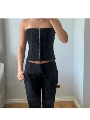 Svart jeanstopp med drajkedja - Säljer en svart tubtopp i denim-look med dragkedja framtill och smockad rygg. Toppen är figurnära och har en cool, trendig vibe. Perfekt att styla med höga byxor eller kjol för en edgy look.