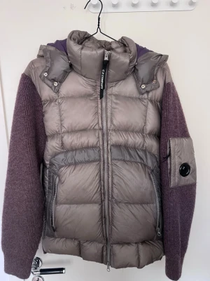 C.P company jacka/cardigan - Unik pufferjacka från C.P. Company med grå quiltad front och huva samt lila stickade ärmar och rygg. Jackan har två stora fickor med dragkedja och en cool detaljficka med lins på ärmen. Perfekt för kalla vinterdagar men även vårdagar och riktigt snygg design.