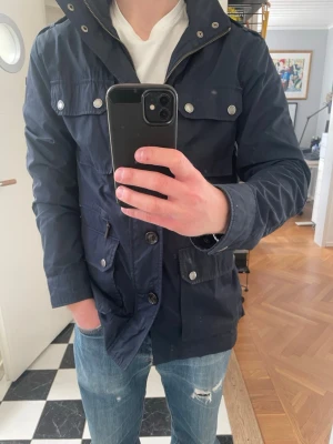 Mörkblå overshirt med fickor - Stilren mörkblå overshirt med fyra framfickor och silverfärgade knappar. Jackan har hög krage, dragkedja och knappstängning. Perfekt för lager-på-lager och passar till jeans eller chinos. Materialet ser ut att vara bomull eller en bomullsblandning.