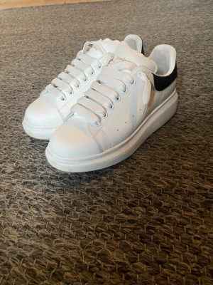 Vita Alexander McQueen sneakers - Snygga vita sneakers från Alexander McQueen med breda vita skosnören och svart detalj på hälen. Skorna har en chunky sula och är tillverkade i läder. Perfekt för dig som gillar stilrena och trendiga sneakers. Lite stora i storlek men d bara att spänna snörena