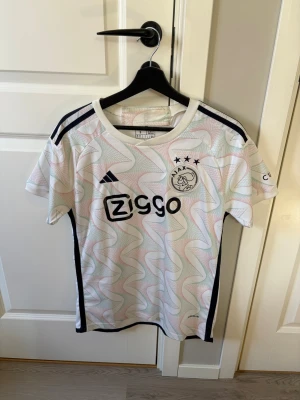Ajax matchtröja Adidas Ziggo vit - Ajax matchtröja i vitt med färgglada grafiska mönster, svarta detaljer och Ziggo-logga på bröstet. Klassisk Adidas-design med tre svarta ränder på axlarna och Ajax klubbmärke med tre stjärnor. Tillverkad i lätt och ventilerande material.