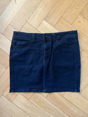 Mörk jeanskjol med fickor - Säljer en mörk jeanskjol med klassiska fickor fram och bak samt hällor för bälte. Kjolen har en rak och kort modell med knapp och dragkedja framtill. Färgen är grå/svart/blå.