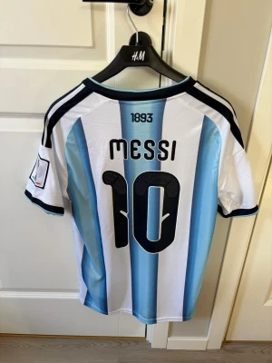 Argentina Messi matchtröja Adidas - Säljer en Argentina matchtröja från Adidas med Messi och nummer 10 på ryggen. Tröjan har klassiska blå och vita vertikala ränder, svarta detaljer på axlar och ärmar samt FIFA- och AFA-emblem på bröstet. Materialet är lätt och andas, perfekt för fotboll.