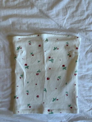 Vit tubtopp med blommigt mönster - Söt vit tubtopp med små röda blommor och gröna blad. Toppen är ribbad och har en bandeau-form som sitter tight runt kroppen. Perfekt för varma dagar och enkel att matcha med jeans eller kjol.