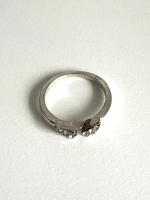 Silverfärgad ring med glittriga stenar - Snygg silverfärgad ring med två bladformade detaljer som är dekorerade med gnistrande kristaller. Ringen har en stilren design som passar perfekt för att lyfta din look. Materialet är metall med syntetiska kristallstenar. 16mm inner-diameter. 