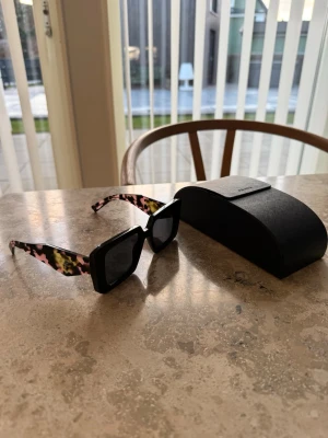 Prada fyrkantiga solglasögon - Säljer ett par statement solglasögon från Prada med fyrkantiga svarta bågar och coola leopardmönstrade skalmar i rosa, svart och gul. Kommer med originalfodral. Perfekta för dig som vill sticka ut och addera färg till din look.