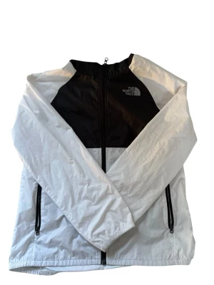 Svartvit vindjacka från The North Face - Snygg svartvit vindjacka från The North Face med dragkedja framtill och två sidofickor med svarta dragkedjor. Jackan har hög krage och luva, samt logga på bröstet. Storlek xl/tg i barn storlek passar runt 160cm