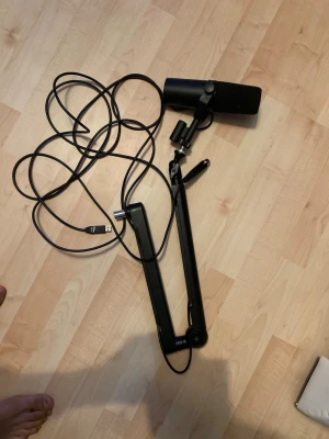 Svart USB-mikrofon med arm - Säljer en svart USB-mikrofon med tillhörande justerbar arm och kabel. Mikrofonen har en cylindrisk form och fäste för enkel montering på skrivbord. Perfekt för streaming, gaming eller podd. Materialet är metall och plast.