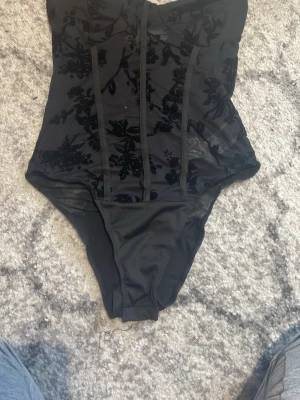 Svart mesh body med sammetmönster - Snygg svart body i mesh med dekorativa sammetmönster och vertikala detaljer framtill. Axelbandslös design och transparenta partier ger en trendig look. Perfekt att styla under en kavaj eller till jeans.