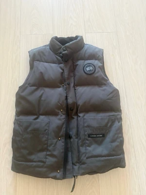 Svart dunväst från Canada Goose - Säljer en svart dunväst från Canada Goose med hög krage, två stora fickor framtill och klassisk logotyp på bröstet. Västen har knappar och dragkedja samt är tillverkad i slitstarkt material med dunfyllning. Perfekt för kalla dagar.